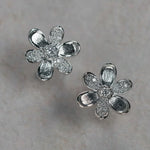 A pair of 18K white gold flower-shaped stud earrings with textured petals and clear diamond centers, displayed on a light gray textured surface.