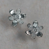 A pair of 18K white gold flower-shaped stud earrings with textured petals and clear diamond centers, displayed on a light gray textured surface.