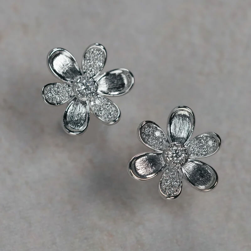 A pair of 18K white gold flower-shaped stud earrings with textured petals and clear diamond centers, displayed on a light gray textured surface.