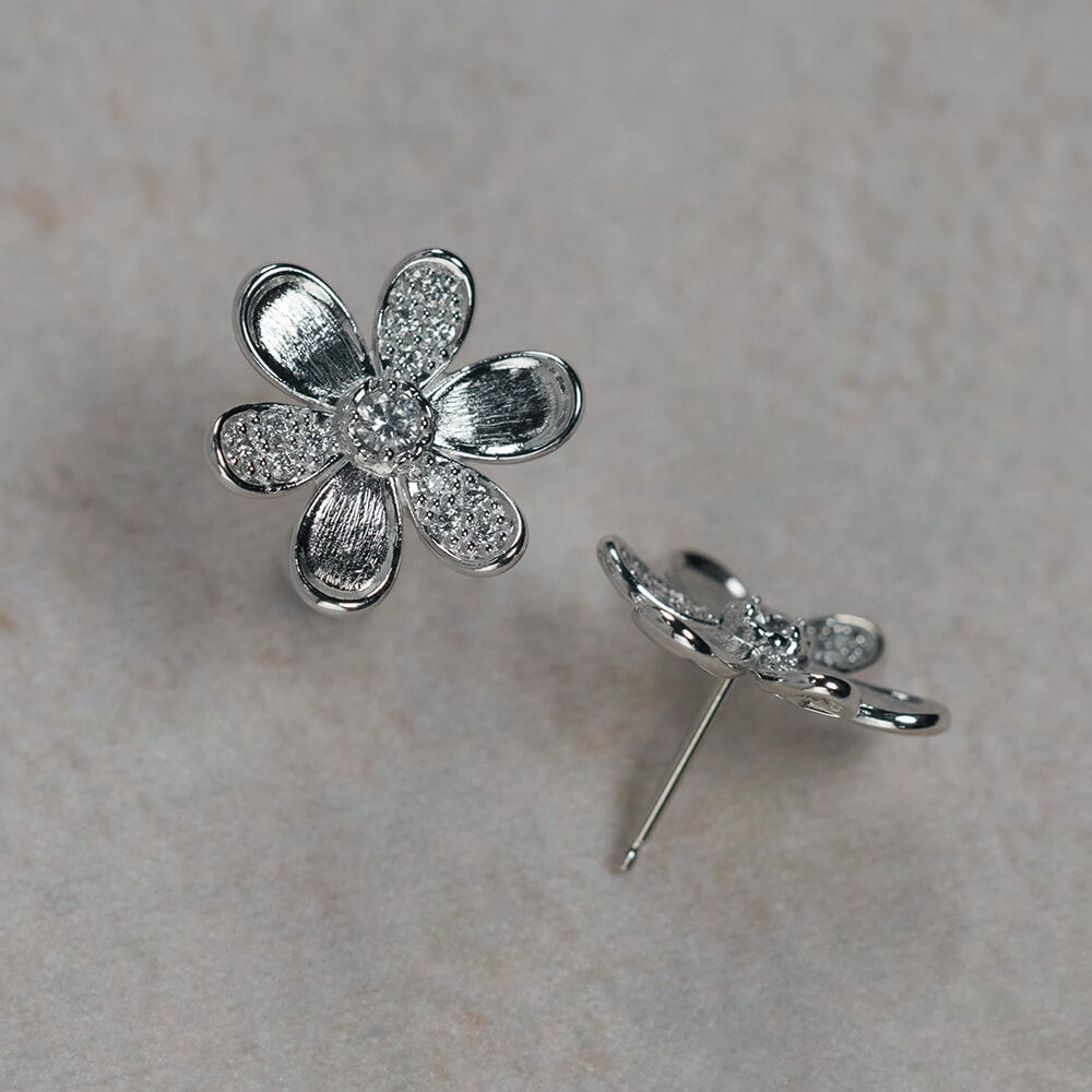A pair of 18K white gold flower stud earrings, one shown from the front with textured and pavé petals, and the other from the side showing the post and backing, set on a light beige textured background.
