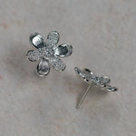 A pair of 18K white gold flower stud earrings, one shown from the front with textured and pavé petals, and the other from the side showing the post and backing, set on a light beige textured background.