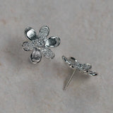 A pair of 18K white gold flower stud earrings, one shown from the front with textured and pavé petals, and the other from the side showing the post and backing, set on a light beige textured background.