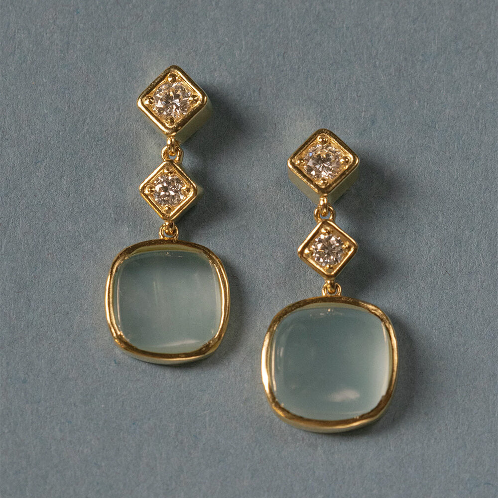 A pair of 18K yellow gold drop earrings with square-cut light blue-green aquamarine stones and small clear diamond accents, displayed on a textured blue-gray background.