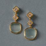 A pair of 18K yellow gold drop earrings with square-cut light blue-green aquamarine stones and small clear diamond accents, displayed on a textured blue-gray background.