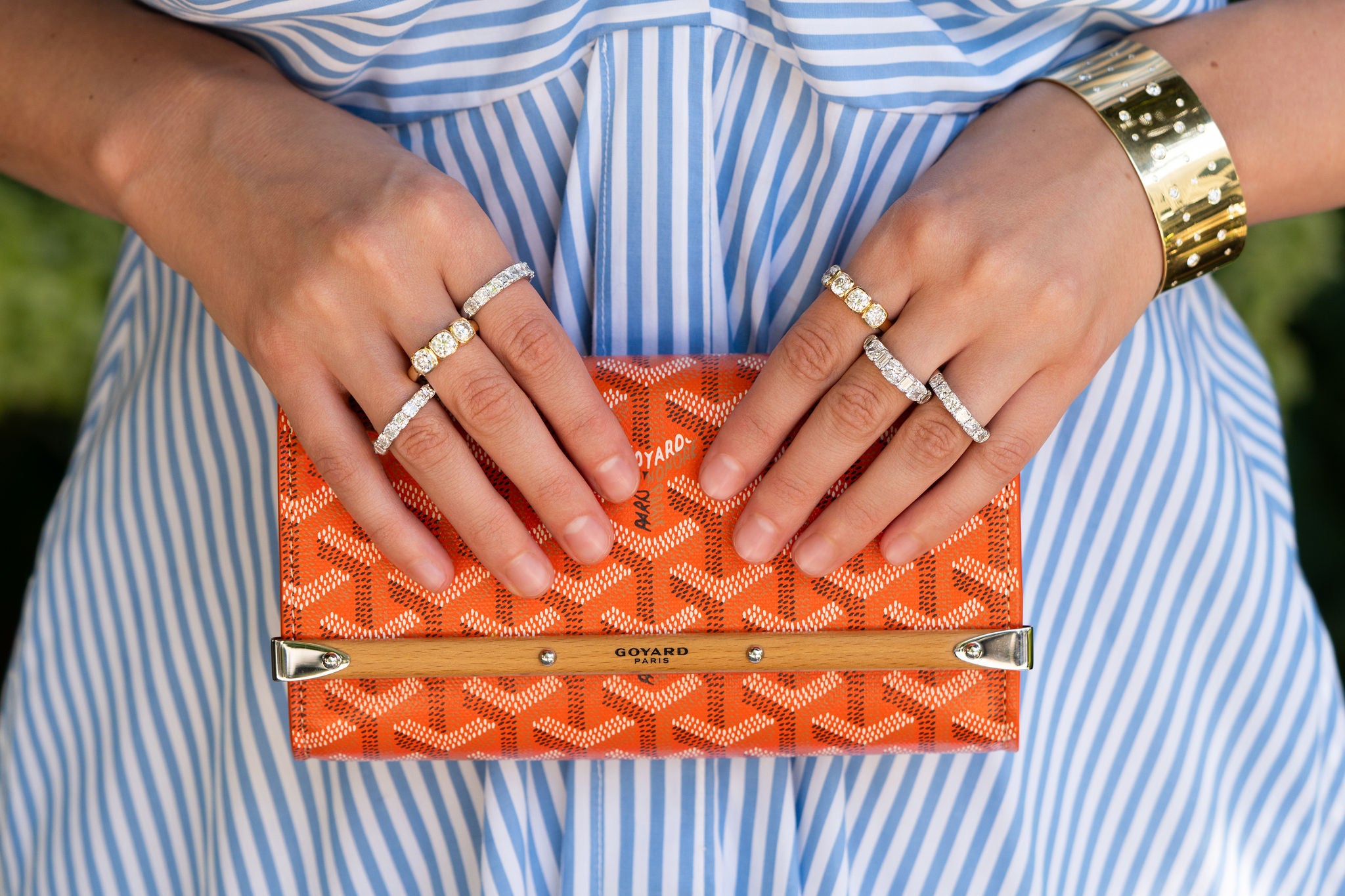 woman wearing multiple diamond band rings and a custom diamond and yellow gold bracelet holding an orange Goyard Paris clutch.