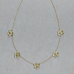 A delicate 18K yellow gold chain necklace featuring five small, spaced-out flower charms with clear faceted diamonds, displayed on a textured light gray background.