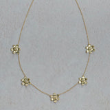 A delicate 18K yellow gold chain necklace featuring five small, spaced-out flower charms with clear faceted diamonds, displayed on a textured light gray background.