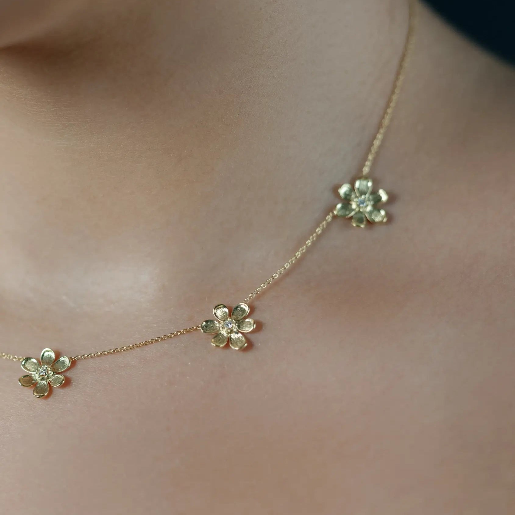 A delicate gold chain necklace with three small flower-shaped charms, shown worn across a person's neck and upper chest.