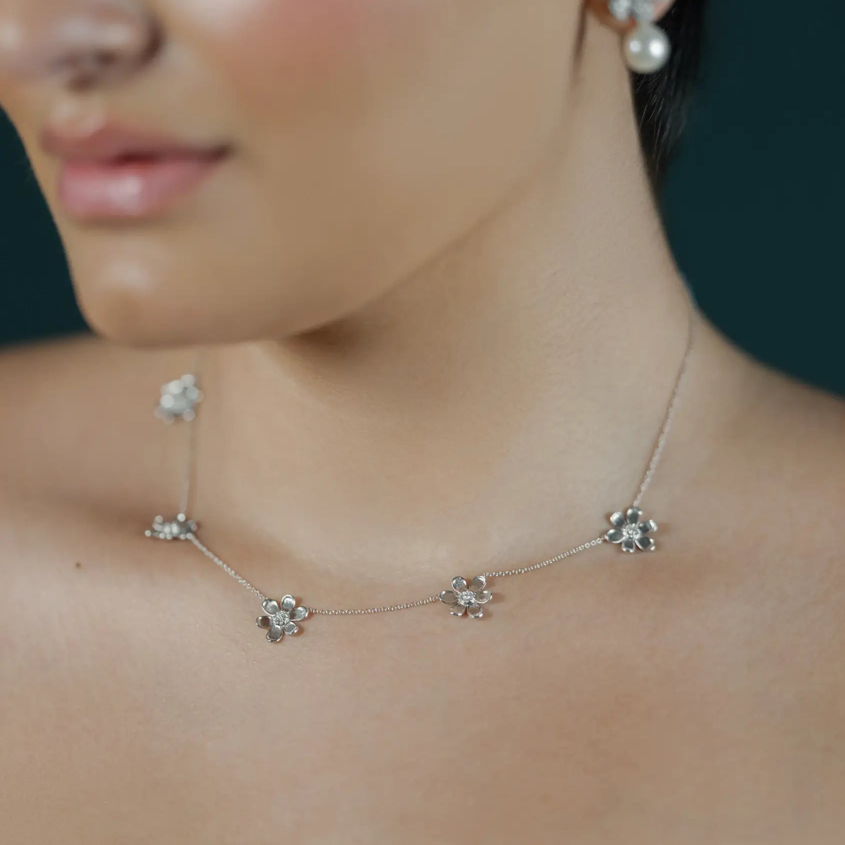 A close-up of a woman wearing a delicate white gold necklace and matching stud earrings with clear floral-like stones and pearl accents against a dark background.