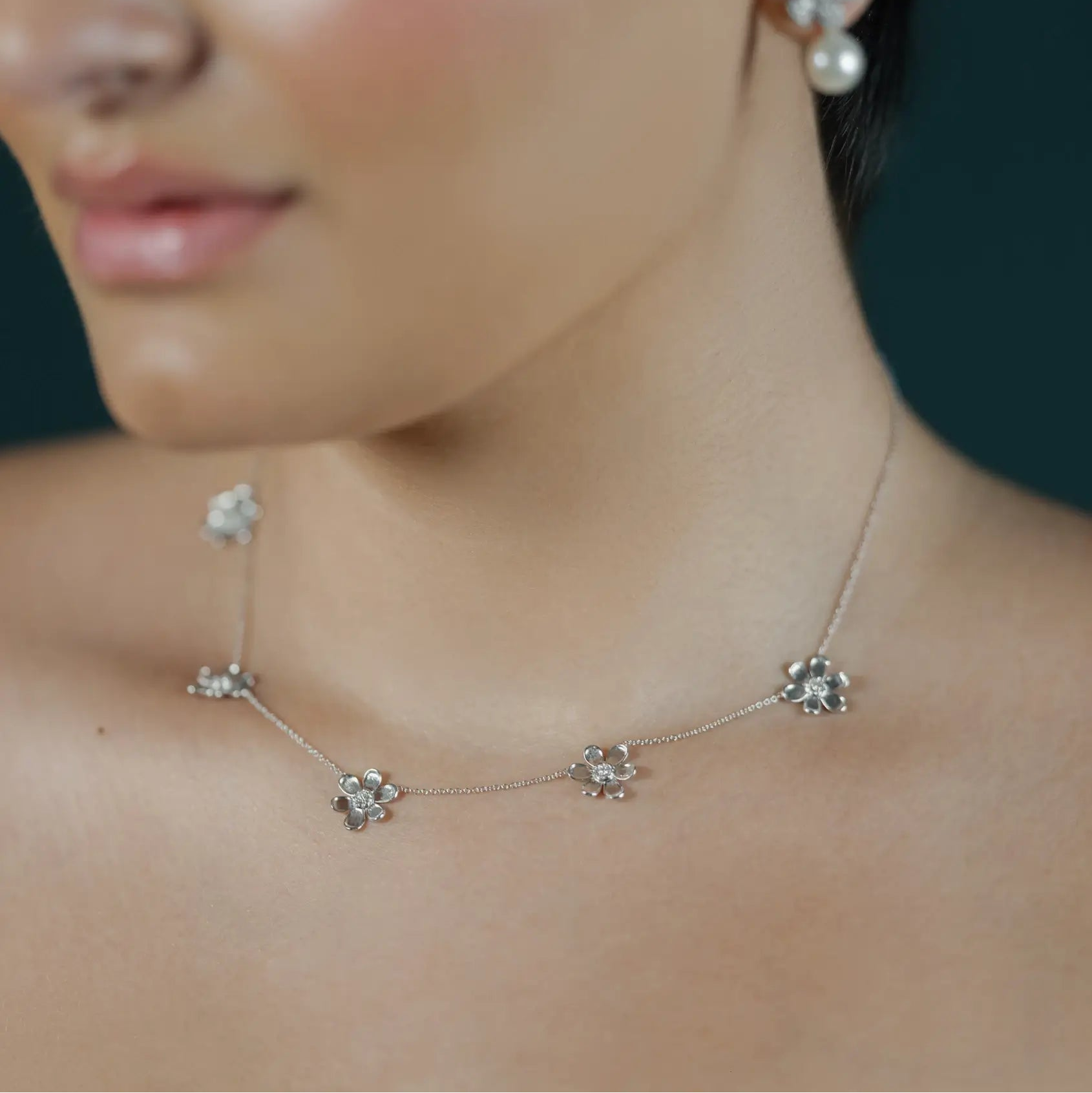 A close-up of a woman wearing a delicate white gold necklace and matching stud earrings with clear floral-like stones and pearl accents against a dark background.
