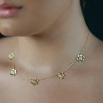 A close-up lifestyle shot of a model wearing a delicate 18K yellow gold chain necklace with five small flower charms, each set with clear diamonds. The model is shown from the side, highlighting the necklace on her neck and collarbone, with a matching diamond stud earring visible.
