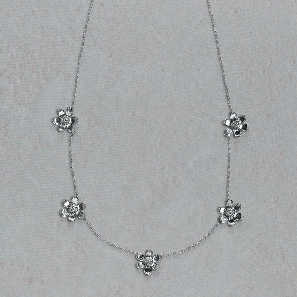 An 18K white gold delicate necklace featuring five small flower-shaped charms spaced along a fine chain, displayed on a light textured neutral background.