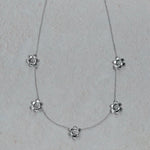 An 18K white gold delicate necklace featuring five small flower-shaped charms spaced along a fine chain, displayed on a light textured neutral background.