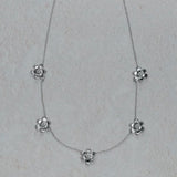 An 18K white gold delicate necklace featuring five small flower-shaped charms spaced along a fine chain, displayed on a light textured neutral background.
