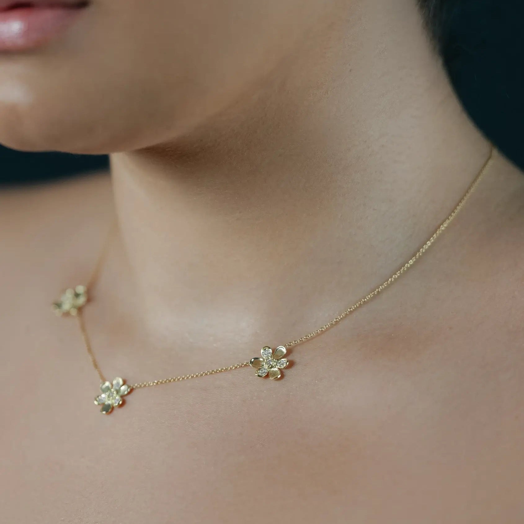 A close-up lifestyle shot of a model wearing a delicate 18K yellow gold chain necklace with three small flower charms with pave diamonds