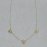 A delicate 18k yellow gold chain necklace featuring three small flower-shaped charms, each with clear diamonds, arranged on a light textured background.