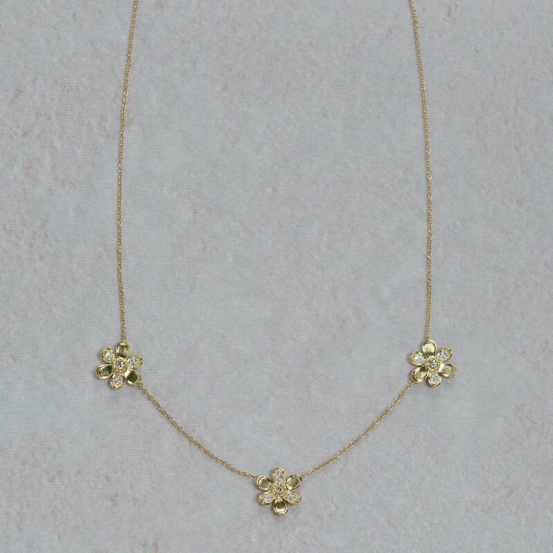 A delicate 18k yellow gold chain necklace featuring three small flower-shaped charms, each with clear diamonds, arranged on a light textured background.