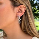 A close-up of a woman's ear wearing a dangling earring made of small clear diamonds arranged in a zigzag pattern set in 18k white gold, with her hair and a blurred outdoor background visible.