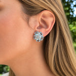 A close-up of a woman's ear wearing an estate 18k white gold flower-shaped stud earring with clear faceted aquamarine gemstones, set against a blurred outdoor background of green foliage.