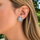 A close-up of a woman's ear wearing an estate 18k white gold flower-shaped stud earring with clear faceted aquamarine gemstones, set against a blurred outdoor background of green foliage.