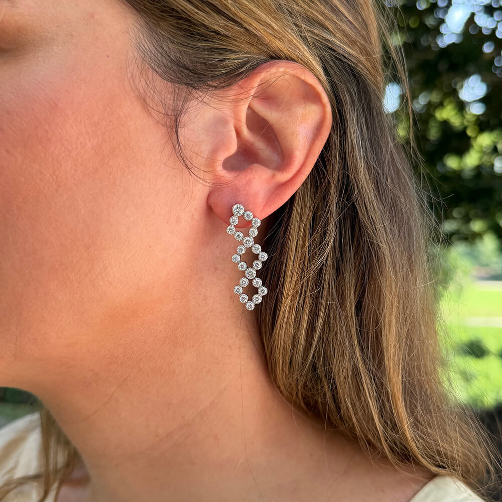 A close-up side profile of a woman wearing a dangling spiral Estate earring made of small clear diamonds on a white gold base, with her ear and hair visible against an outdoor blurred background.