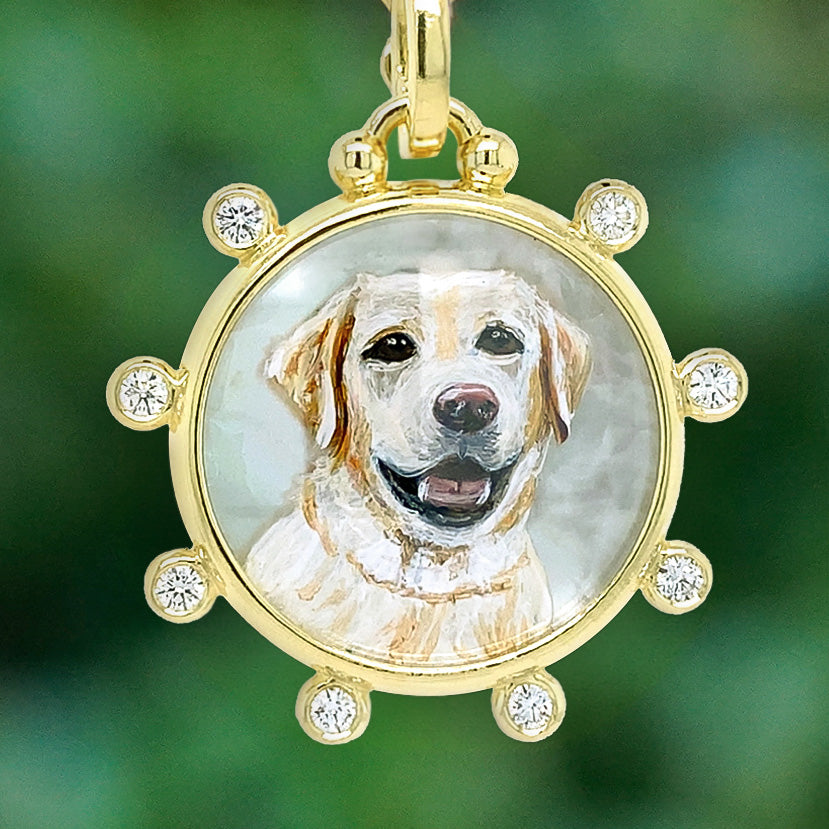A round gold pendant charm featuring a hand-painted portrait of a yellow Labrador Retriever, surrounded by a gold frame with small clear rhinestones, set against a soft green blurred background.