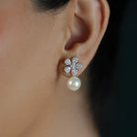 A close-up of a woman's ear wearing an 18K white gold flower stud earring with clear pavé stones and a dangling white pearl.