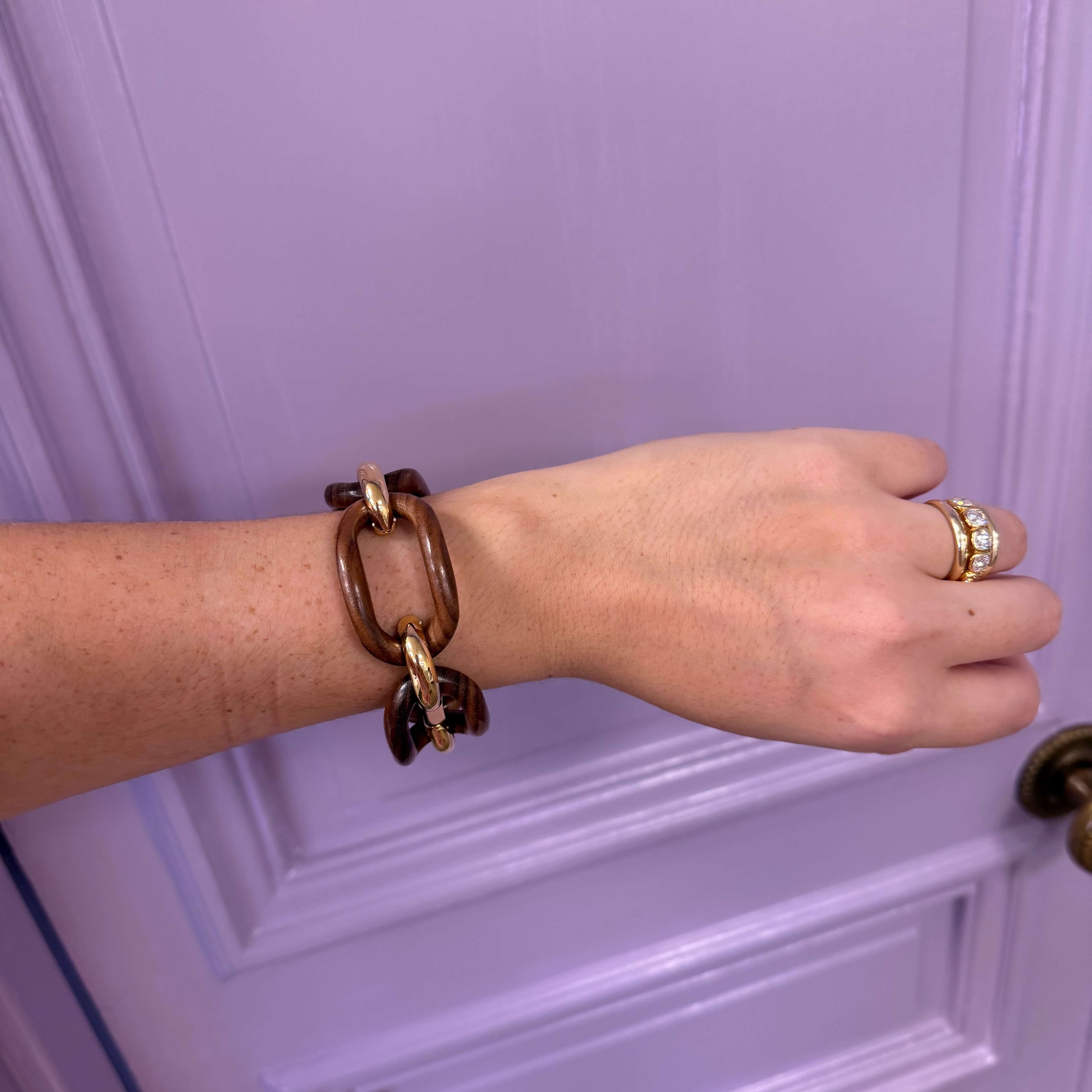 Hand wearing a brown chain bracelet and gold ring against a purple door.