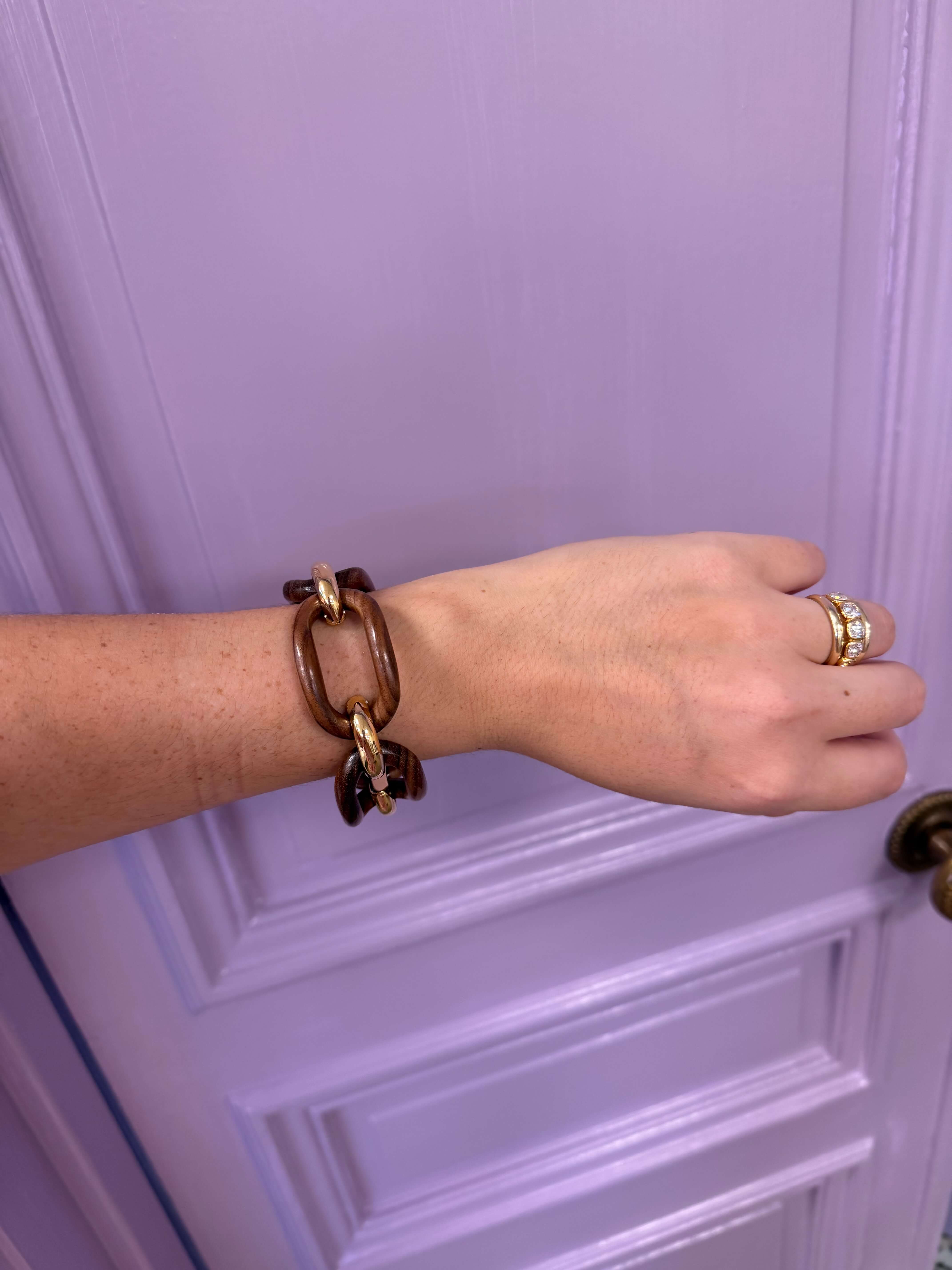 Hand wearing a brown chain bracelet and gold ring against a purple door.