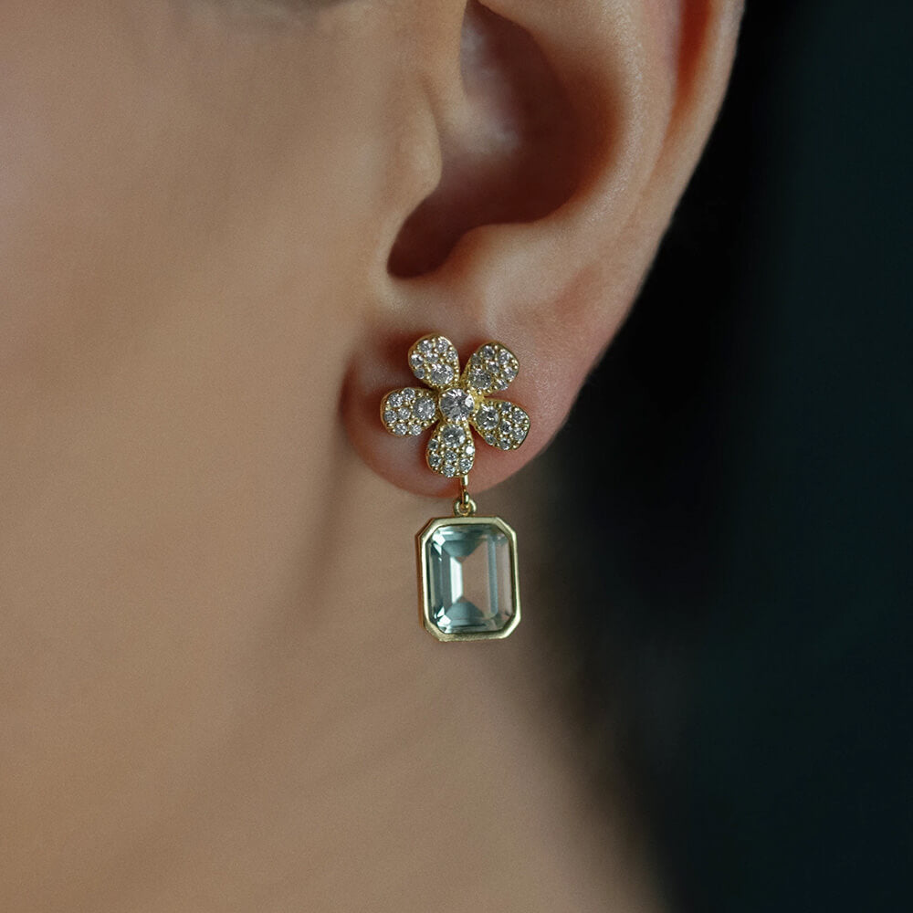 A close-up of a woman's ear wearing an 18K yellow gold flower stud earring with pavé-set clear diamonds and a dangling rectangular emerald-cut green anethyst gemstone.