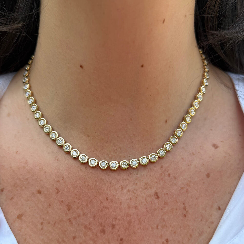 A close-up of a 14K yellow gold tennis necklace with small round clear diamonds, worn on a person's neck and upper chest.