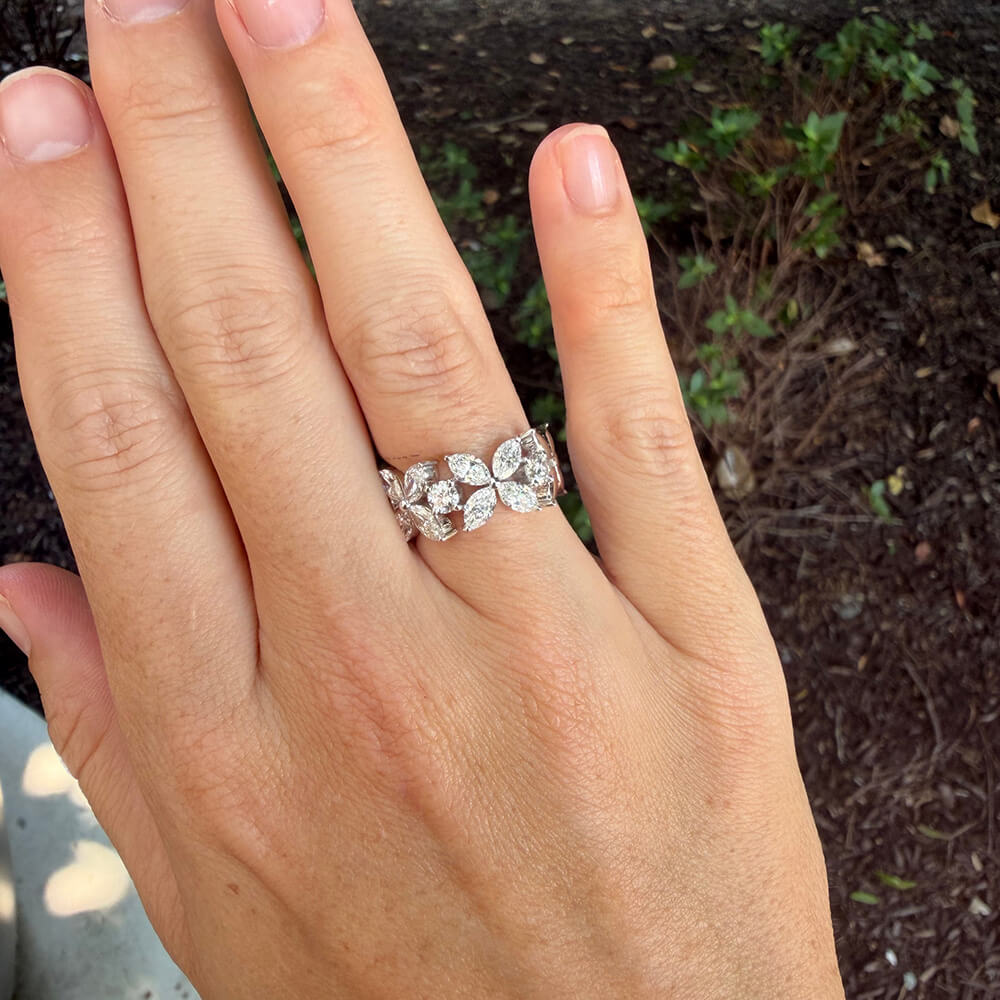 An 18K white gold ring with marquise-cut and round diamonds arranged in a leafy pattern, shown worn on a woman's hand against a blurred outdoor background.