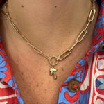 A close-up of a woman's neck and chest wearing a 14K yellow gold chain necklace with a small mushroom charm, paired with a red and blue patterned button-up shirt.