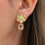 A close-up of a woman's ear wearing a gold flower-shaped stud earring with small clear diamonds and a dangling rectangular morganite gemstone earring jacket