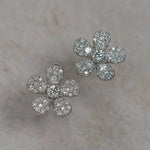 A pair of 18K white gold flower-shaped stud earrings paved with clear diamonds, displayed on a light beige textured surface.