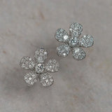 A pair of 18K white gold flower-shaped stud earrings paved with clear diamonds, displayed on a light beige textured surface.