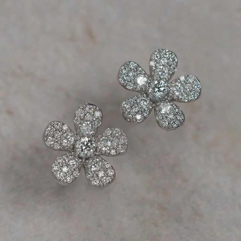 A pair of 18K white gold flower-shaped stud earrings paved with clear diamonds, displayed on a light beige textured surface.