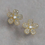 A pair of 18K yellow gold flower-shaped stud earrings paved with clear diamonds, displayed on a textured light gray surface.