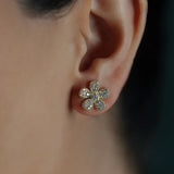 A close-up of a woman's ear wearing a flower-shaped stud earring with five petals covered in pave diamonds in an 18K yellow gold setting.