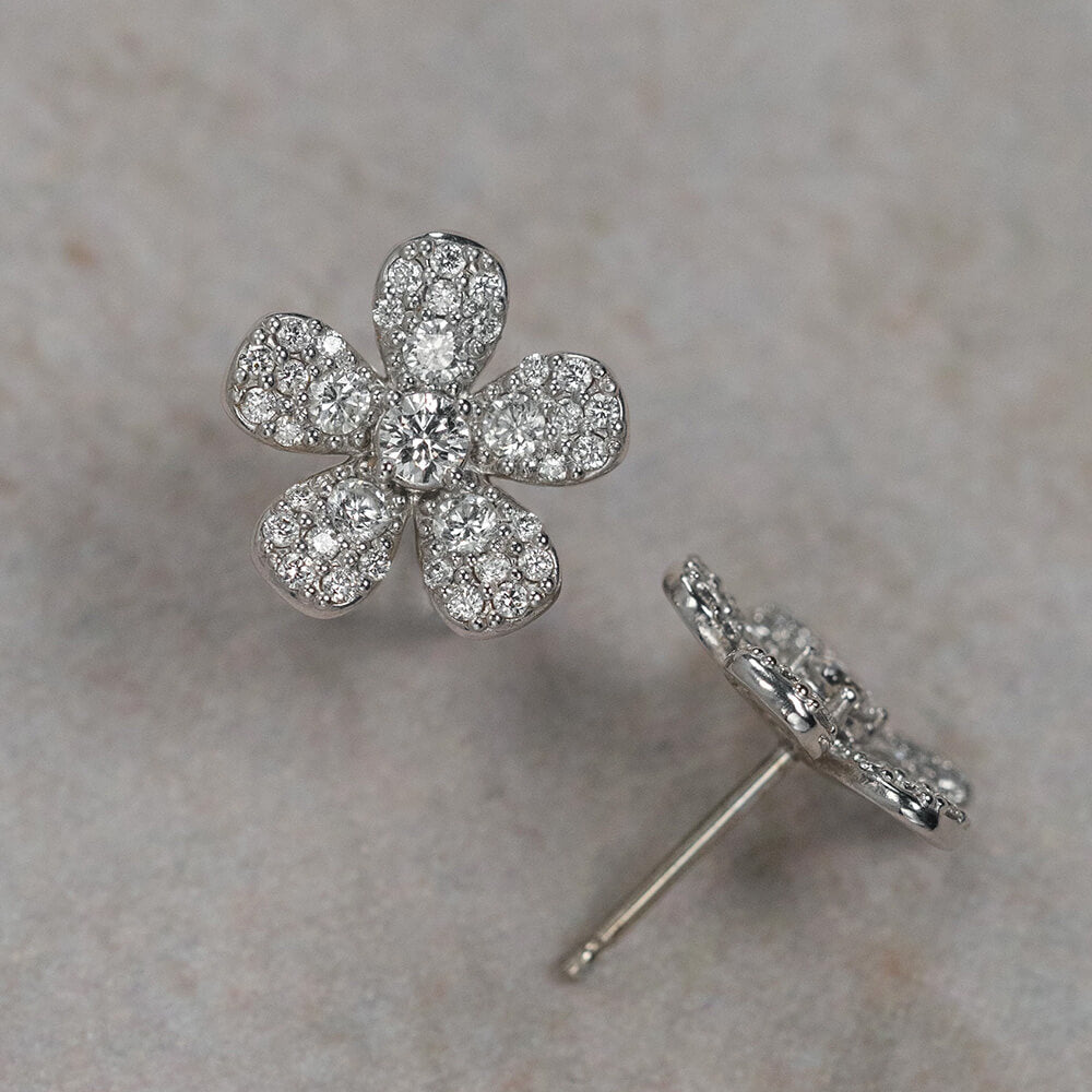 A pair of 18K white gold flower-shaped stud earrings paved with clear diamonds, with one earring shown from the front and the other from the side/back, resting on a textured beige surface.