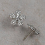 A pair of 18K white gold flower-shaped stud earrings paved with clear diamonds, with one earring shown from the front and the other from the side/back, resting on a textured beige surface.
