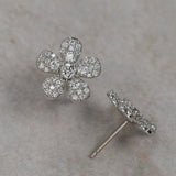 A pair of 18K white gold flower-shaped stud earrings paved with clear diamonds, with one earring shown from the front and the other from the side/back, resting on a textured beige surface.