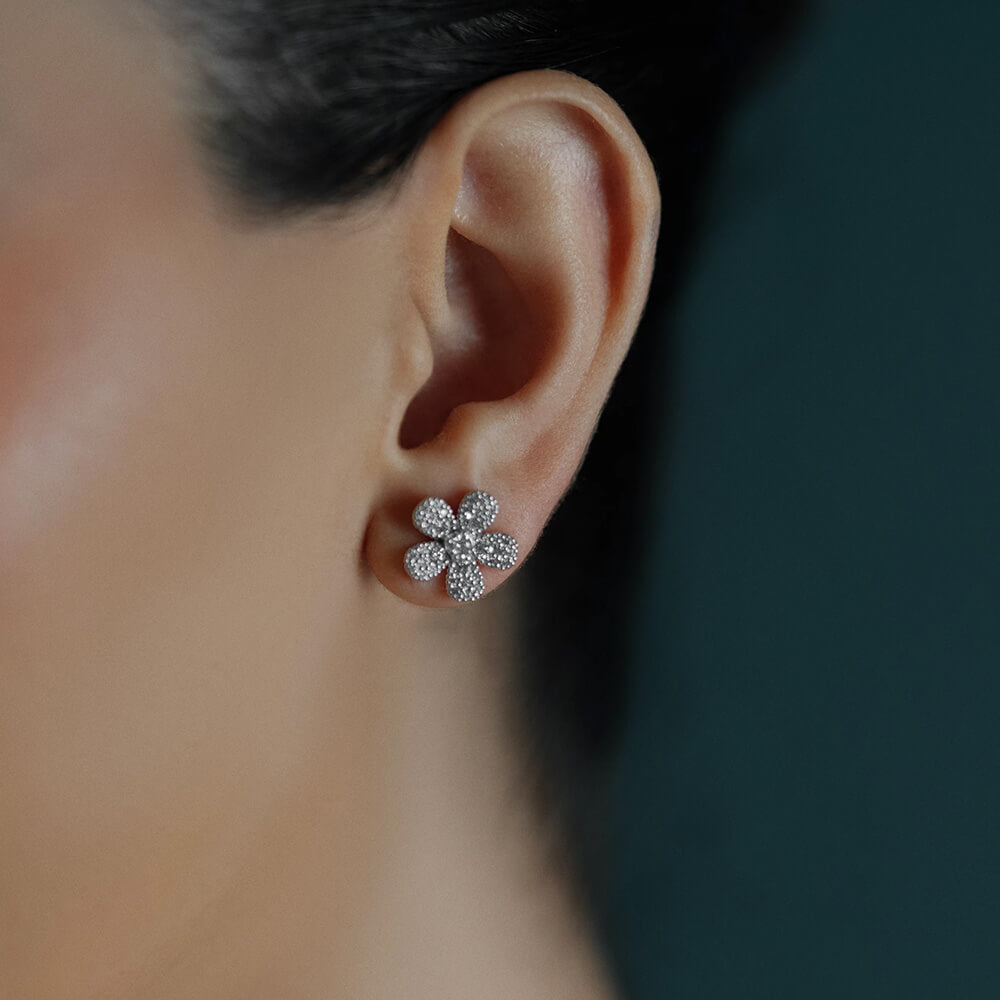 A close-up of a woman's ear wearing a flower-shaped stud earring set with clear diamonds in an 18K white gold metal, against a dark teal background.
