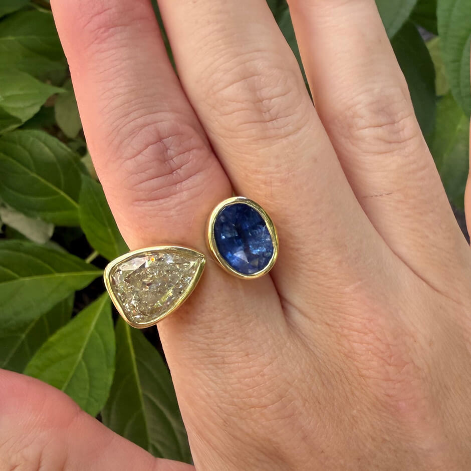 A close-up of a hand wearing an 18K yellow gold ring with a triangular clear diamond and an oval deep blue sapphire, set against a background of green leaves.