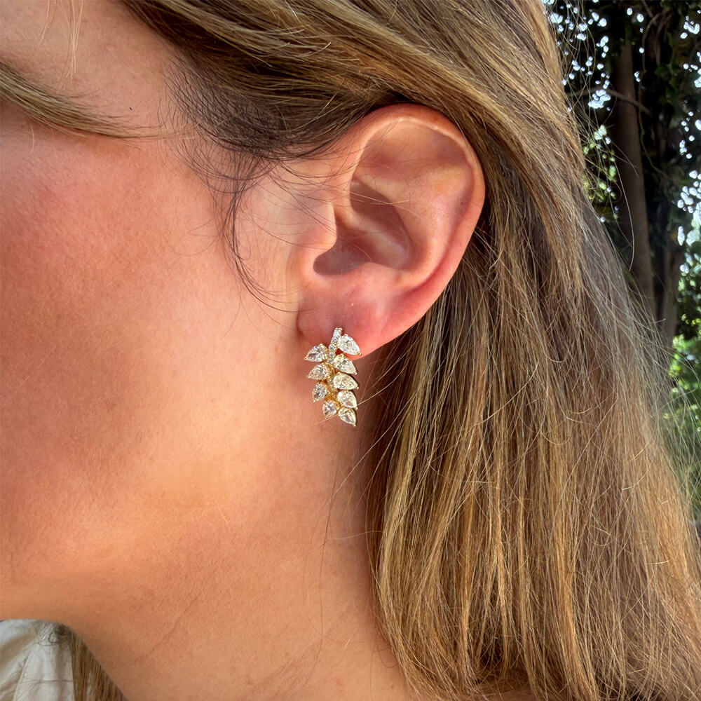 A close-up of a woman's ear wearing a 18K yellow gold leaf-shaped stud earring set with clear pear-shaped diamonds.