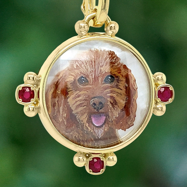 A gold pendant locket with a custom hand-painted portrait of a brown dog under a clear glass cover, accented with four red gemstones on a blurred green background.