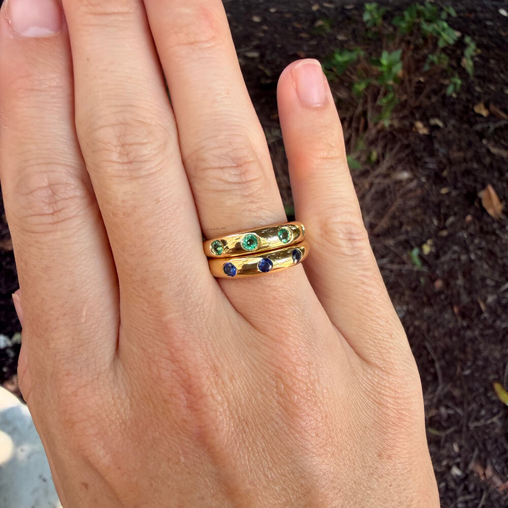 A close-up of a hand wearing two stacked 14K yellow gold rings, each set with small round emerald green and sapphire blue gemstones, against an outdoor background of dark soil and plants.