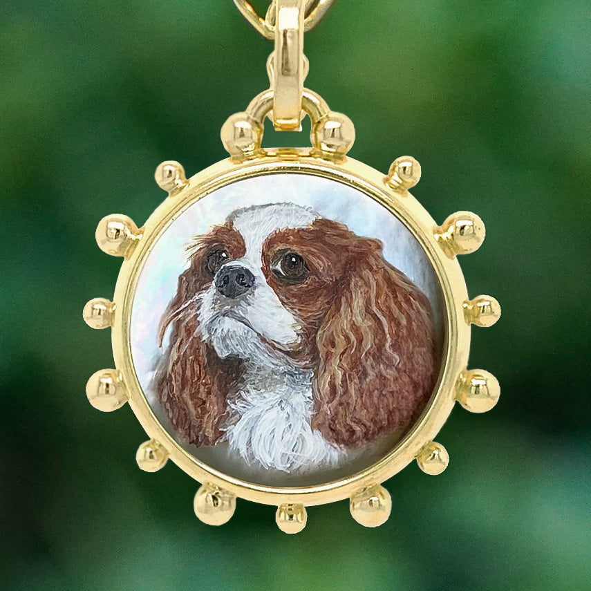 A round gold pendant necklace featuring a hand-painted portrait of a brown and white Cavalier King Charles Spaniel dog, set against a soft, blurred green background.
