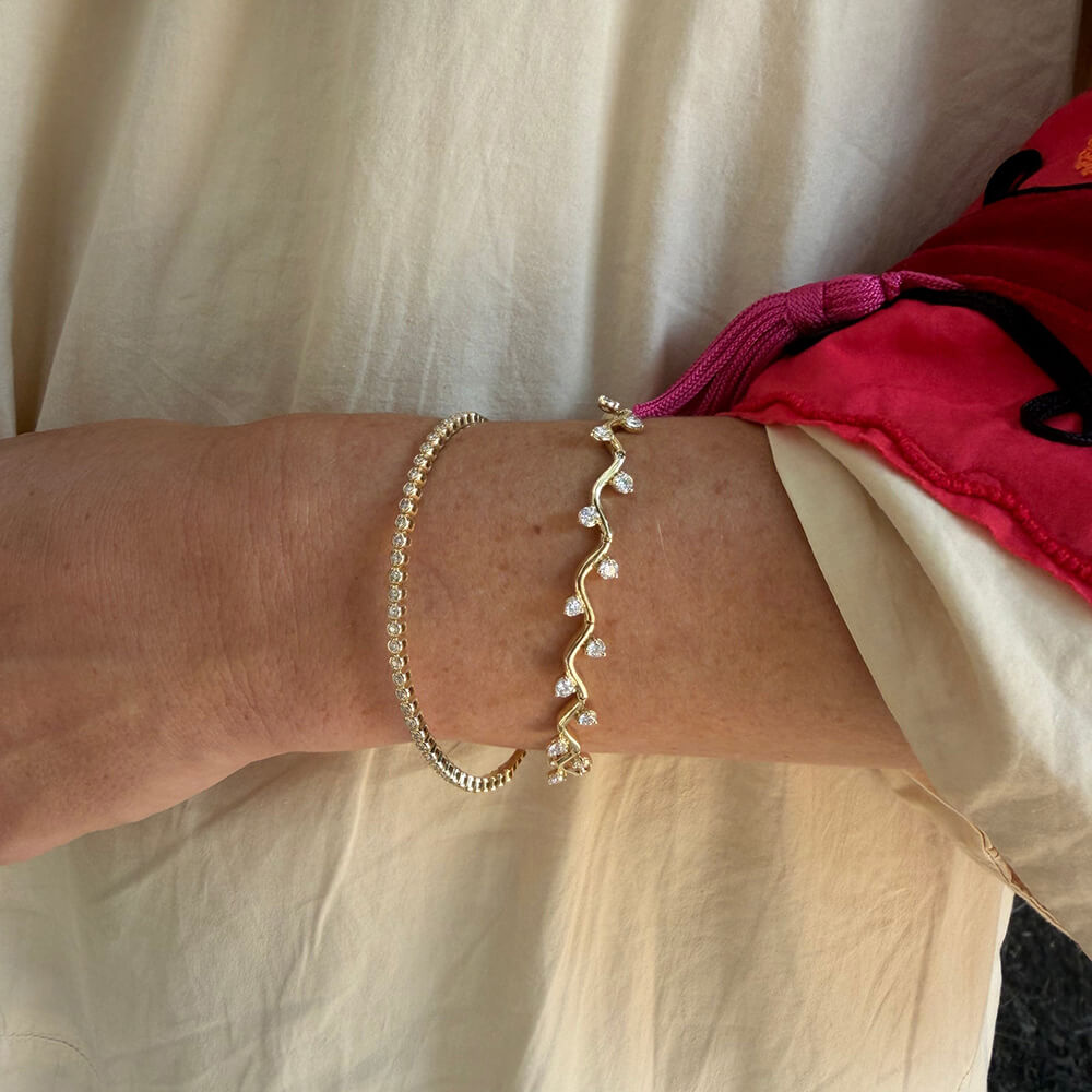A close-up of a woman's wrist wearing two delicate bracelets: one gold bangle with a wavy line of clear diamonds and another diamond bangle.