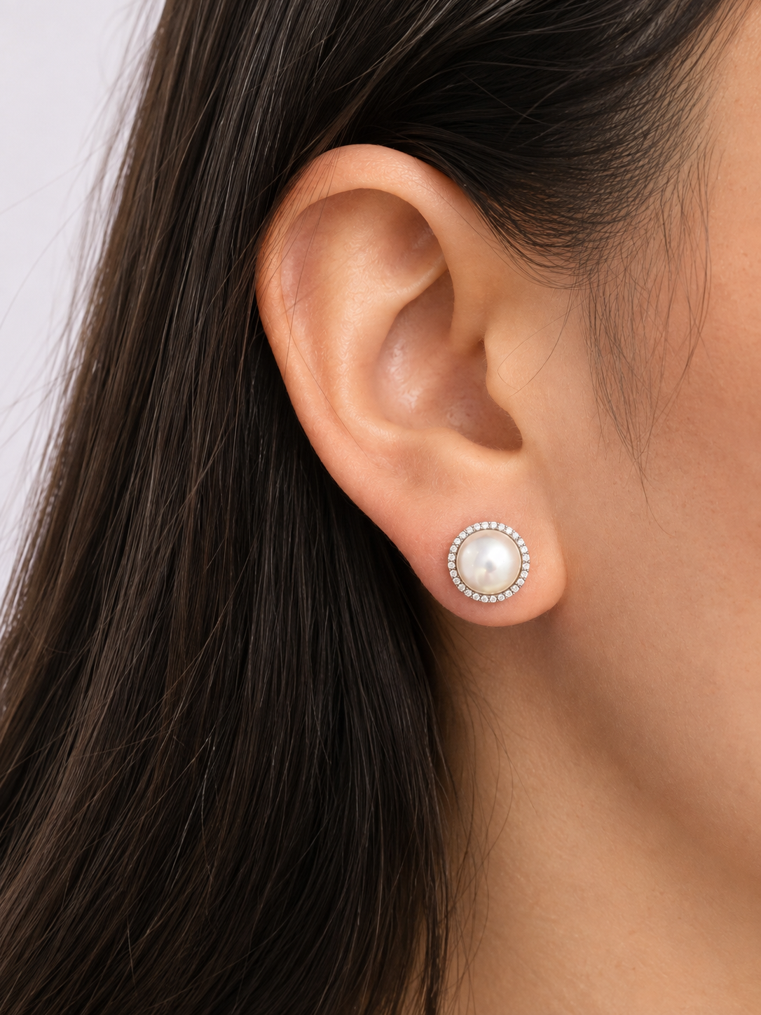 A close-up of a model's ear wearing a stud earring with a central white pearl surrounded by a halo of diamonds